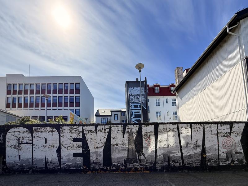 A wall with a mural that says 'GREYKJAVIK'. The lettering contains the skyline of Reykjavík. The sky is blue and the sun is shining.