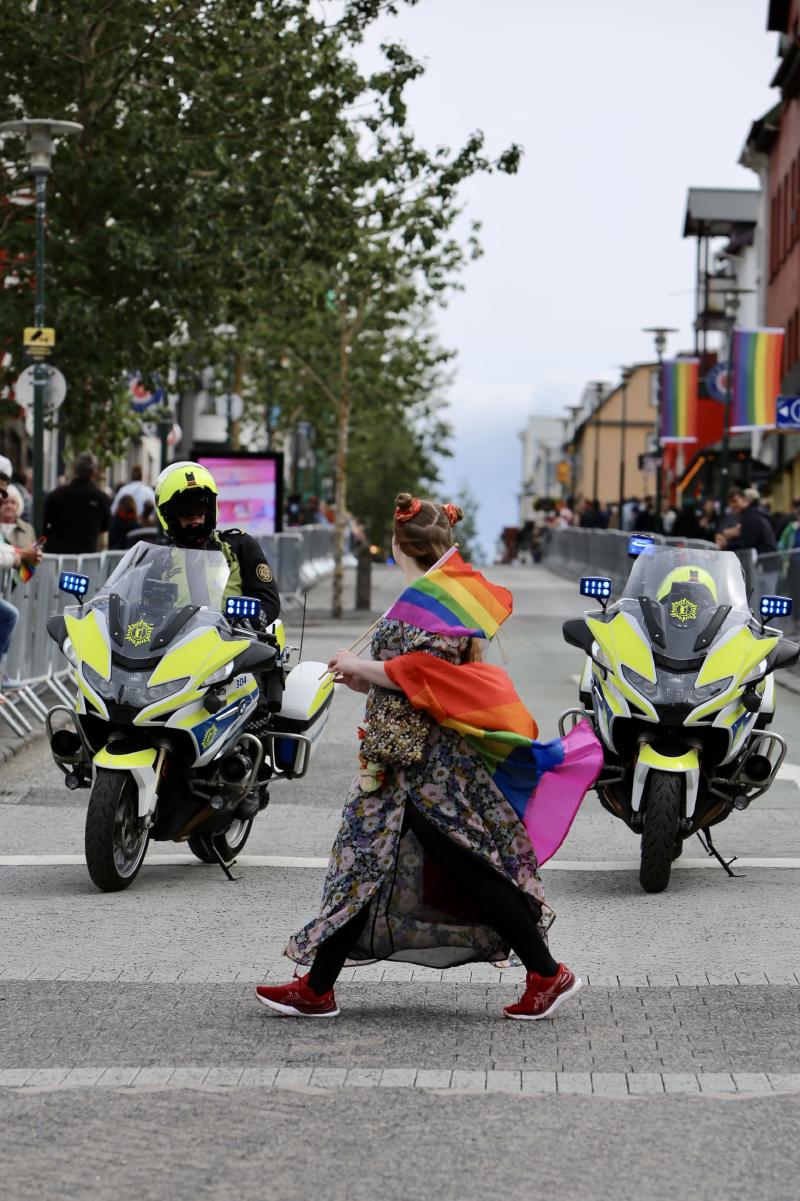 A woman wearing a rainbow flag walks between two police motorcycles in front of a crowd of people.