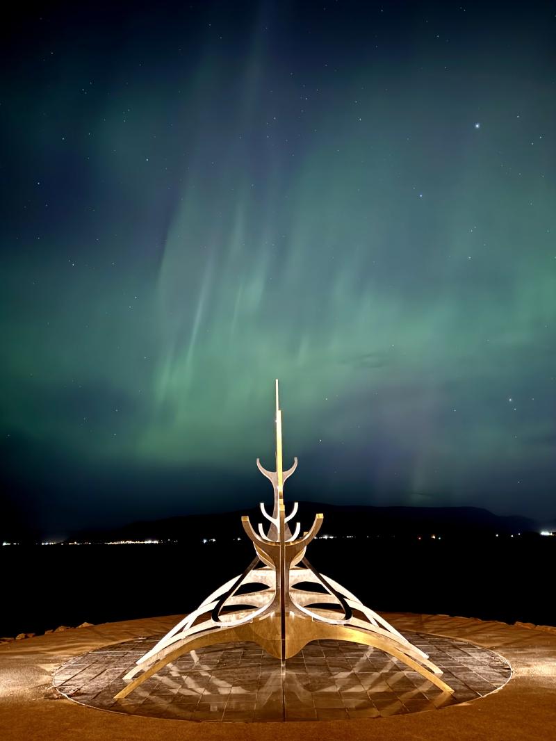 A ship sculpture sits on a circular stone platform under a starry sky with green auroras.